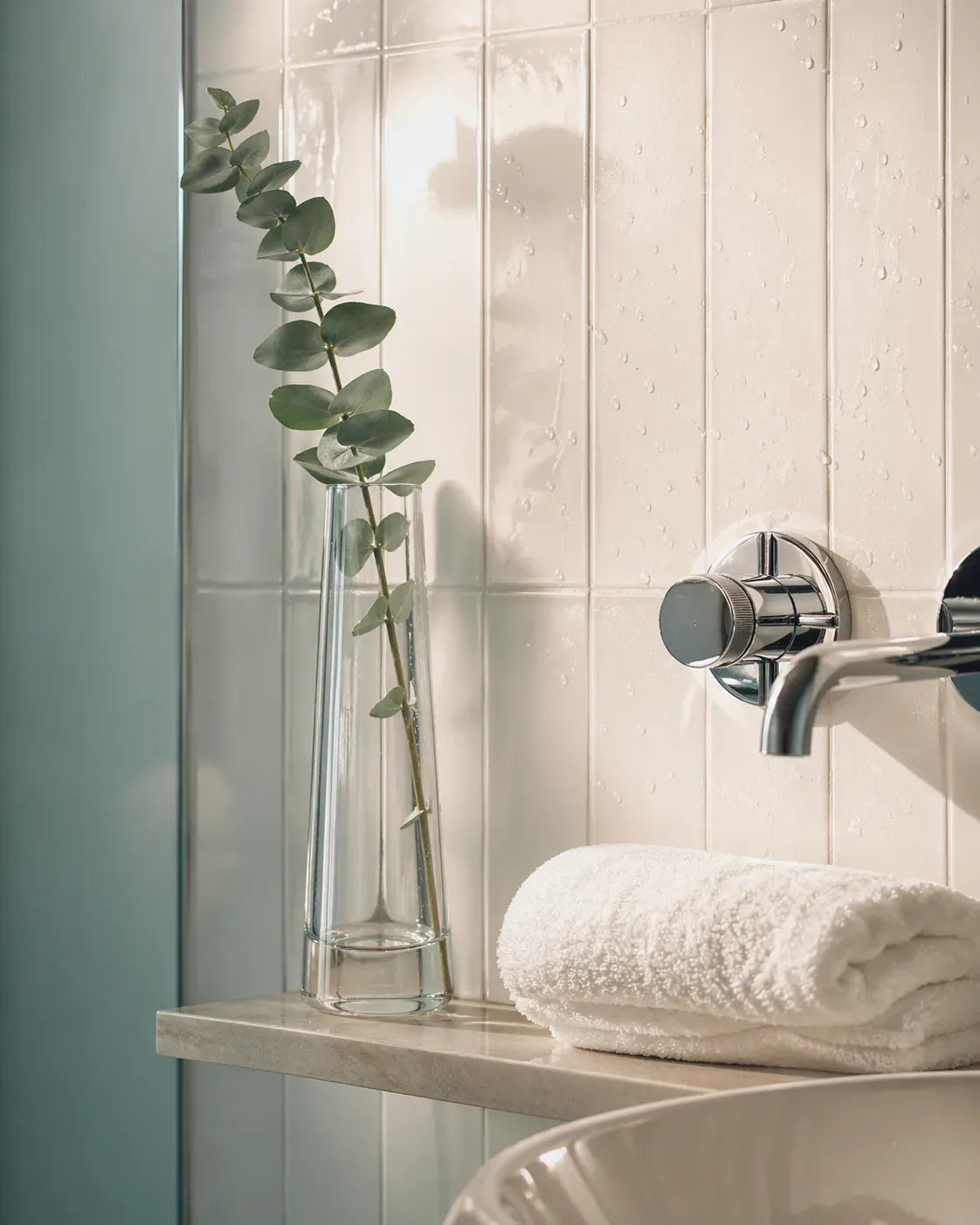 A pristine white subway-tiled bathroom corner with polished chrome fixtures, water beading on tile, and a single eucalyptus stem in a clear vase